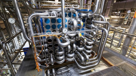 In einer Produktionsstätte stehen verschiedene große Maschinen mit Rohren und Verbindungen.