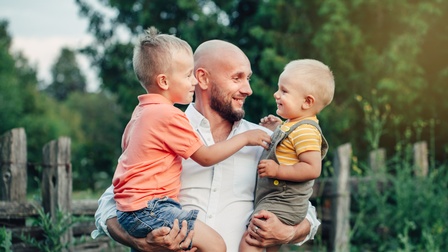 Lächelnde Person mit Bart hält zwei Kinder am Arm, im Hintergrund verschwommen Holzzaun und Pflanzen