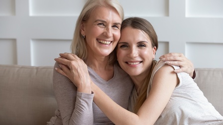 Zwei Personen sitzen auf einem Sofa. Die rechte Person hat ihren Kopf auf der linken Schulter der anderen Person. Die linke Person hat ihre linke Hand auf der linken Schulter der anderen Person. Im Hintergrund ist eine weiße Holzvertäfelung