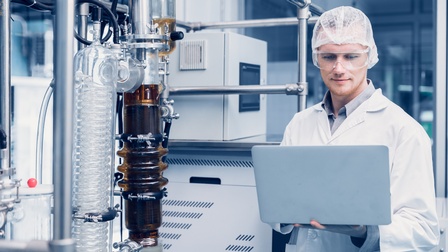 Person mit Haarnetz und Schutzbrillen blickt auf aufgeklappten Laptop, neben bei Extraktionsmaschine zur Distillation