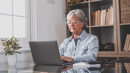 Person mit grauen Haaren und Brille sitzt an Tisch und blickt auf Laptop