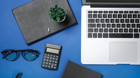 Draufsicht auf einen Arbeitsplatz mit aufgeklapptem Laptop, einer kleinen Pflanze und weiteren Arbeitsutensilien.