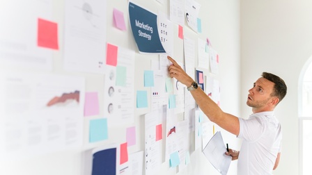 Person steht vor Wand voller Notizblätter und Ideenpapieren und deutet auf ein Blatt mit dem Schriftzug Marketing Strategy