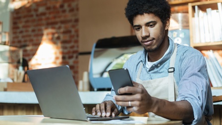 Eine Person mit Schürze sitzt an einem kleinen, runden Tisch und blickt auf ein Handy, das sie hält. Auf dem Tisch steht ein aufgeklappter Laptop. Im Hintergrund ist ein Tresen