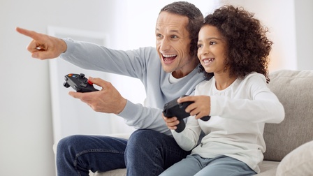 Erwachsene Person sitzt mit einem Kind auf einer Couch und spielt Playstation