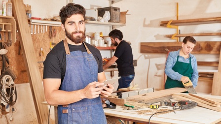 Person mit Bart in blauer Schürze Tablet haltend steht in Werkstatt im Fokus, im Hintergrund Werkbank mit Werkzeugen, Regale und zwei weitere Personen bei Holzabreiten