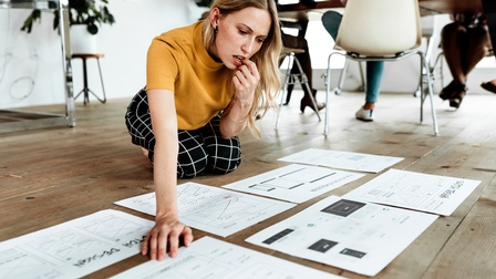Person mit blonden mittellangen Haaren sitzt am Boden und betrachtet ausgedruckte Unterlagen für Web-Design während weitere Personen an einem Tisch im Hintergrund sitzen