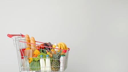 Einkaufswagen vor grauer Wand gefüllt mit Gemüse, Obst, Brot, Milch und Wasser