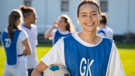 Portrait einer lächelnden jugendlichen Person in blauem Trikot mit weißer Aufschrift GK, unter dem Arm Fußball haltend, im Hintergrund verschwommen weitere Personen in Fußballdressen