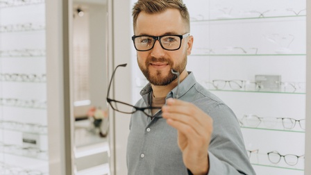 Sanft lächelnde Person mit Brillen hält Brille in der Hand, im Hintergrund Regale voller Brillen