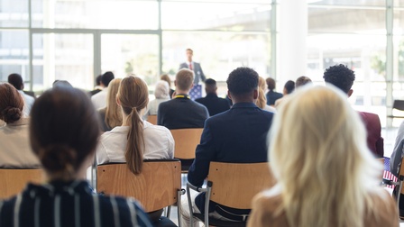 Rückenansicht vieler Menschen, die in Sesselreihen in einem großen Raum mit Fensterwänden sitzen. Im Hintergrund steht eine Person an einem Rednerpult, die zum Publikum spricht