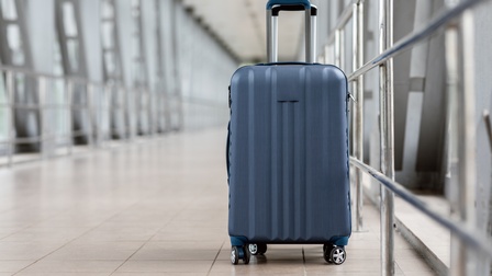 Ein kleiner, blauer Koffer mit Rollen und ausgefahrenem Griff steht neben einem metallenen Geländer auf Fliesen in einem hellen Gang