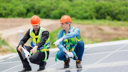 Zwei Personen in gelben Warnwesten mit orangen Schutzhelmen hocken auf Trittfläche auf Dach mit Solarpanelen und deuten auf bestimmte Stelle, im Hintergrund verschwommen belaubt Büsche