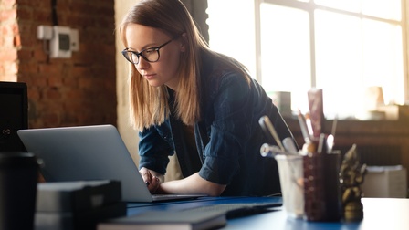 Person, die sich über einen Schreibtisch beugt und auf einen geöffneten Laptop blickt. Die Hände stützen sich auf der Tischplatte ab. Auf dem Schreibtisch stehen unter anderem ein Stiftebehälter und weitere Arbeitsutensilien