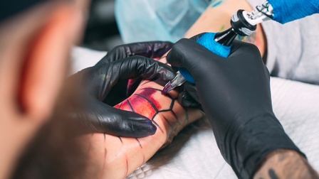 Nahaufnahme von der Erstellung eines Tattoos mit bunten Farben, Person trägt schwarze Handschuhe und tätowiert die Hand einer Person