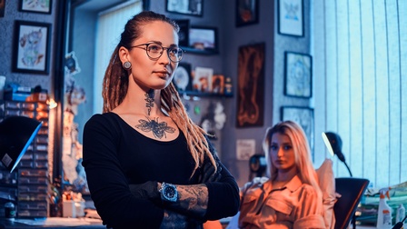 Porträt einer Person mit Brille, einer gepiercten Lippen, Tätowierungen und verschränkten Armen. Hinter ihr sitzt eine Person in einem Stuhl, die in die Kamera blickt. Neben ihr ist eine Stehlampe