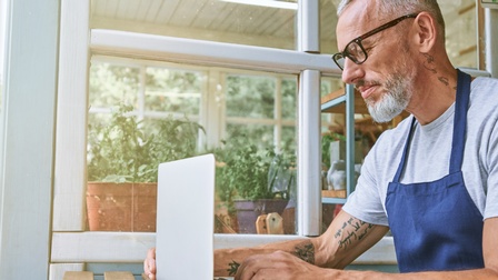 Person mit kurzen grauen Haaren, Bart, Tattoos und Brille trägt eine blaue Schürze und sitzt bei einem Gartentisch mit einem aufgeklappten Laptop, dahinter zeigt sich ein Fenster eines Glashauses mit Pflanzen