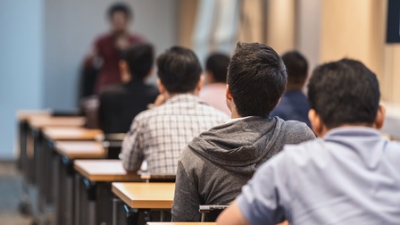 Rückenansicht von mehreren Personen, die hintereinander einzeln an einem Tisch sitzen. Ihnen gegenüber steht eine Person