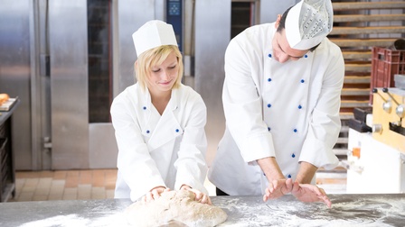Zwei Personen in weißen Oberteilen mit Mützen stehen nebeneinander hinter einem metallenen Tisch. Vor der linken Person liegt ein großer Teig, den sie knetet sowie Mehl. Die andere Person blickt auf den Teig und ahmt mit den Händen eine Knetbewegung nach.