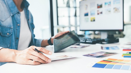 Person sitzt vor einem Schreibtisch mit Unterlagen, Farbkarten, Stiften und hält ein Tablet in der Hand, im Hintergrund steht ein großer Monitor