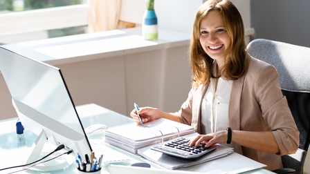 Lächelnde Person sitzt an einem Schreibtisch mit Laptop und geht eine Mappe mit Unterlagen durch, während mit einem Taschenrechner gerechnet wird