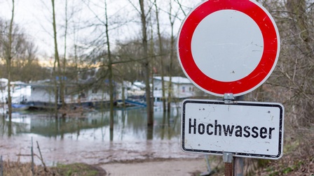 In der rechten Bildhälfte ist ein Verkehrsschild, auf dem Hochwasser steht. Darüber ist ein weißer Kreis mit einem roten Rand. Im Hintergrund führt ein Weg zu einer Fläche mit Häusern und Bäumen, die unter Wasser steht