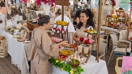 Zwei Personen stehen an einem Marktstand und sprechen miteinander. Auf mehrstöckigen Regalen sind handgefertigte Produkte, darunter Kerzen, Seifen und dekorative Artikel, ausgestellt. Im Hintergrund befinden sich weitere Marktstände sowie mehrere Personen