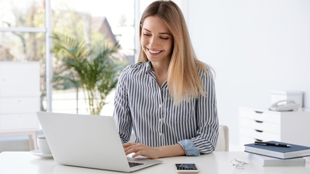 Eine Frau sitzt in einem hellen Büro mit großflächigem Fenster vor einem Laptop. Dieser ist auf einem Schreibtisch platziert, rechts davon ist eine Tasse, links ein Smartphone, eine Brille, zwei Bücher übereinander und ein Kugelschreiber