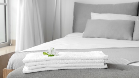 Weiße Handtücher liegen auf einer grauen Kleiderablage vor einem weiß-grau bezogenem Bett