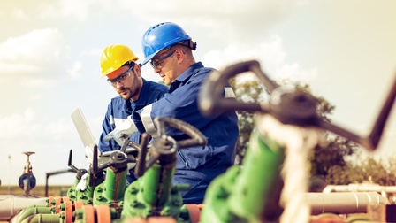 Personen mit Arbeitskleidung, Schutzhelme und Schutzbrillen halten einen Laptop in den Händen und blicken darauf während sie bei ei Gasrohren stehen