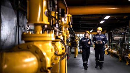 Personen in Schutzkleidung mit gelben Schutzhelmen stehen in einer Raffinerie und führen Qualitätskontrollen mit einem Tablet durch
