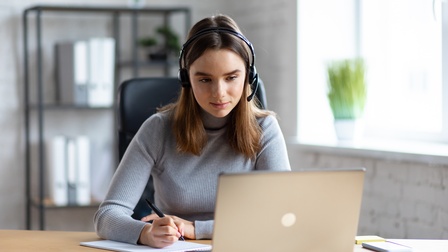 Frontale Aufnahme einer Person mit offenem, braunem Haar und Headset, die an einem Tisch sitzt. Mit der rechten Hand hält sie einen Stift. Darunter liegt ein Notizblock. Daneben steht ein aufgeklappter Laptop