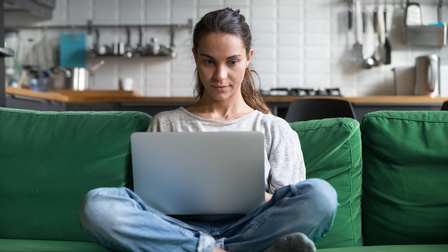 Person sitzt im Schneidersitz auf grünem Sofa und blickt auf aufgeklapptes Notebook am Schoß, im Hintergrund verschwommen Küchenzeile