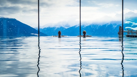 Zwei Personen lehnen am Beckenrand eines Hallenbades. Hinter ihnen ist ein Panoramafenster. Dort sind man schneebedeckte mit Wolken verhangene Berge