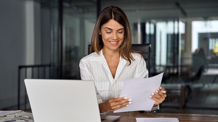 Person mit braunen schulterlangen Haaren und gestreifter weißer Bluse sitzt an einem Schreibtisch in einem gläsernen Büro und begutachtet freudig Unterlagen, während ein aufgeklappter Laptop neben ihr steht