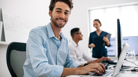 Person sitzt an einem Schreibtisch und blickt lächelnd in die Kamera und während am Laptop gearbeitet wird, im Hintergrund unterhalten sich weitere Personen 