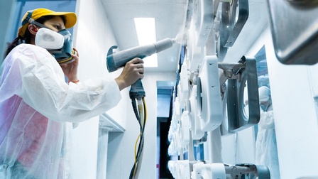 Person mit weißem Schutzanzug und Atemmaske sprüht Farbe auf ein Objekt in einer Werkstätte