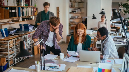 Fünf Personen in einem Büro. Drei Personen beschäftigen sich gemeinsam mit Unterlagen und einem Laptop und sitzen bzw. stehen an einem Tisch. Im Hintergrund befinden sich zwei weitere Personen