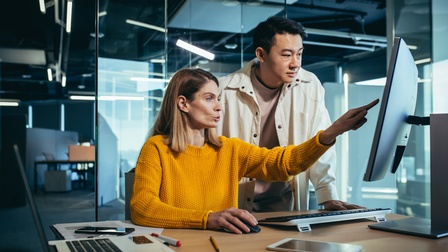 Zwei Personen befinden sich an einem Arbeitsplatz in einem Büro mit Glaswänden. Eine Person sitzt vor einem Bildschirm und bedient eine Computermaus. Die zweite Person steht daneben. Beide richten ihren Blick auf den Monitor