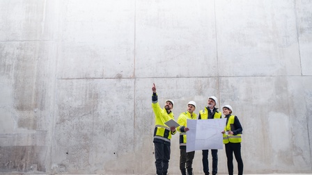 Vier Personen in gelben Warnwesten mit weißen Schutzhelmen stehen vor Betonwand und blicken nach oben in Fingerzeigrichtung einer der Personen, zwei der Personen halten großen Papierplan