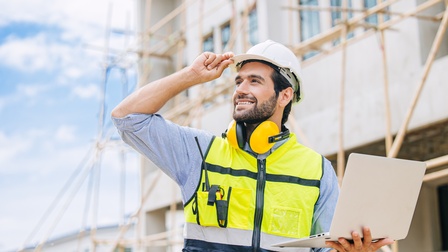 Frontale Aufnahme einer Person mit Helm, Sicherheitsweste, Gehörschutz um den Hals und einem Walkie Talkie in der Brusttasche, die auf einer Baustelle steht und einen Laptop hält. Die Person blickt nach links oben