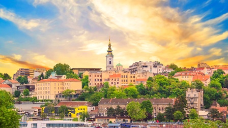 Panoramaaufnahme der Stadt Belgrad mit historischen und modernen Gebäuden. In der Mitte ragt ein Kirchturm in die Höhe. Im Vordergrund ist ein Fluss mit Booten. Am Himmel sind Wolken