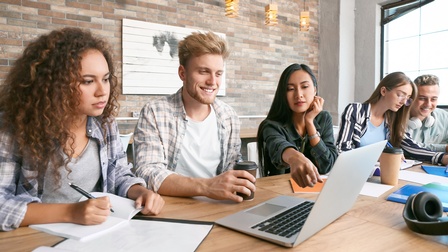 Fünf Personen sitzen nebeneinander an einem langen Tisch in zwei Kleingruppen. Drei der Personen blicken auf einen aufgeklappten Laptop. Die anderen beiden Personen notieren etwas in einem Notizbuch