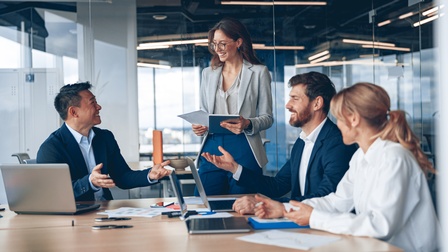 Mehrere Personen in Businesskleidung sitzen bei einem Besprechungstisch in einem modernen hellen, aus Glas abgetrennten Meetingraum