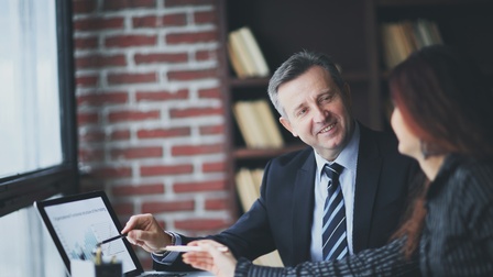 Seitliche Aufnahme von zwei Personen, die nebeneinander an einem Schreibtisch sitzen. Vor ihnen steht ein aufgeklappter Laptop mit einer Statistik, auf den beide deuten. Die Personen blicken sich an