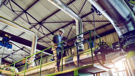 Person in Businesskleidung und Schutzhelm steht auf einer Plattform und blickt auf ein Tablet in einem Innenraum eines Kraftwerkes