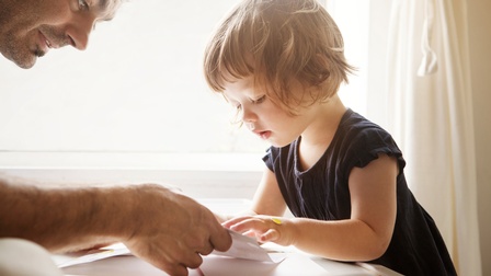 Kleinkind mit blauem Kleid spielt gemeinsam mit einer erwachsenen Person an einem Kinderspieltisch