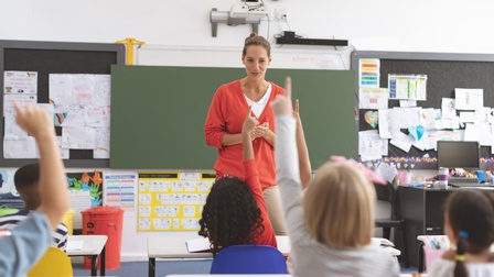Eine erwachsene Person unterrichtet Kinder in einem Klassenzimmer. Die Kinder sitzen an Tischen. Einige heben gerade die Hand. Im Hintergrund sind eine Tafel und zahlreiche Unterrichtsmaterialien zu sehen.