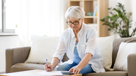 Person mittleren Alters mit Brille sitzt auf einer Couch in einem Wohnraum und stützt sich auf einen Beistelltisch ab während sie dabei auf Unterlagen blickt und einen Stift sowie einen Taschenrechner bedient