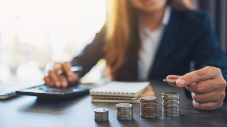 Auf einem Tisch sind vier, unterschiedlich hohe Münzstapel, Notizblöcke sowie ein Taschenrechner, auf dem eine Person tippt. Die Person legt auf den Münzstapel ganz rechts eine weitere Münze.